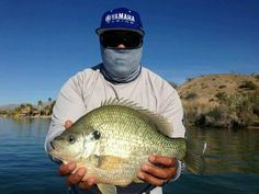 a man holding a large fish while wearing a face mask on the side of a body of water