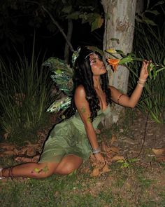 a woman dressed as a fairy sitting on the ground next to a tree and holding a flower in her hand