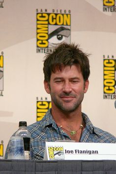 a man sitting in front of a microphone at a press room table with two bottled water bottles