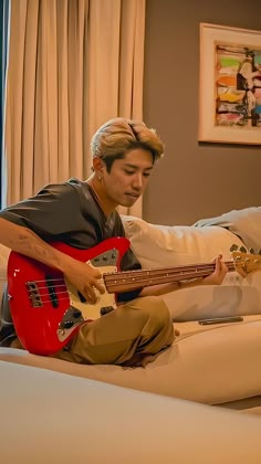 a man sitting on top of a bed while holding a red bass guitar in his hand