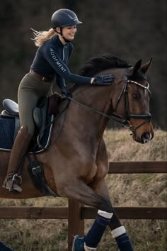 a woman riding on the back of a brown horse next to a wooden fence and grass