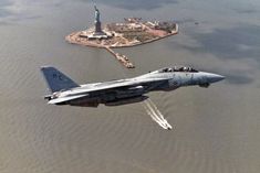 an air force jet flying over a body of water near a statue of liberty in the distance