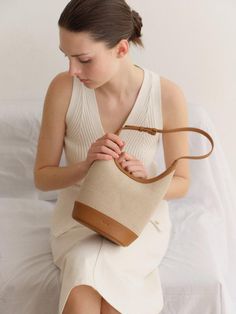 a woman sitting on top of a bed holding a beige handbag in her right hand