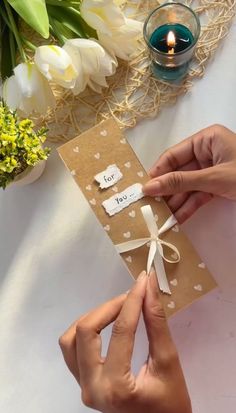 two hands holding a brown card with white hearts on it next to a candle and flowers