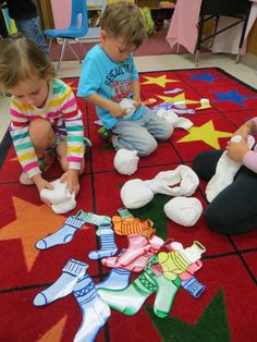 three children sitting on the floor playing with their shoes and socks in a room full of toys