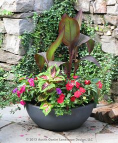 a planter filled with lots of colorful flowers next to a stone wall and shrubbery