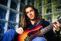 a man sitting in front of a glass block wall holding a guitar and looking at the camera