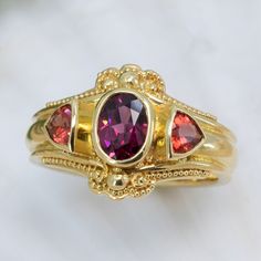 Wrapped in a luxurious setting of 18K granulated gold, our elegant Garnet & Sunstone ring offers a captivating display of gorgeous fall colors. The striking centerpiece is a beautiful rhodolite garnet stone, set off by two sunset-hued sunstones that sparkle with subtle warmth. This exquisite piece will add timeless sophistication to any wardrobe. 18KY granulated ring with 7x5mm rhodolite garnet and 2- 4mm trillion cut sunstones. The granulation on runs part way down the band resulting in a band Gold Heirloom Ruby Ring With Gemstone Accents, Heirloom Gold Ruby Ring With Gemstone Accents, Luxury Garnet Rings With Accent Stones, Gold Garnet Birthstone Ring With Center Stone, Gold Ruby Birthstone Ring With Gemstone Accents, Yellow Gold Ruby Ring With Tourmaline Center Stone, Heirloom Ruby Ring With Gemstone Accents, Elegant 22k Gold Ruby Ring, Gold Garnet Multi-stone Ring