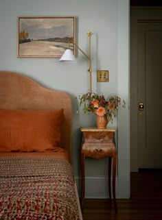 a bed with an orange blanket next to a painting on the wall and a small wooden table