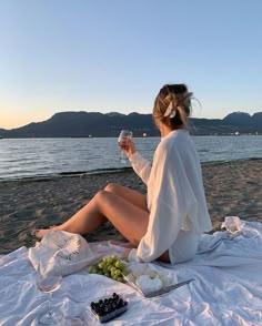 a woman sitting on the beach holding a glass of wine and eating grapes next to water