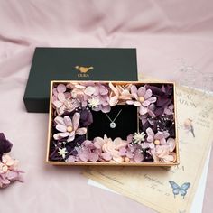 an open box with flowers and a necklace in it on a pink cloth covered surface