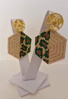 These elegant earrings feature a unique hexagonal design, handcrafted with delicate Miyuki beads in beige, gold, and green tones that create a geometric pattern. The contrast between the colors brings a bold and sophisticated design to life. The top of the earrings has a round detail made of bronze filigree plated in 24-karat gold, adding a luxurious and artisanal touch. Length: 5 cm Width : 4 cm Each piece is unique and reflects the delicacy and precision of the artisanal technique. Please feel free to reach out to us with special requests. Custom orders could take up to 2 months Thank you Handmade Geometric Gold Earrings, Gold Geometric Beaded Earrings As A Gift, Gold Geometric Beaded Earrings For Gift, Geometric Beaded Gold Earrings, Gold Beaded Geometric Earrings, Geometric Gold Beaded Earrings, Gold Geometric Beaded Earrings, Geometric Beaded Gold Jewelry, Gold Beaded Geometric Jewelry