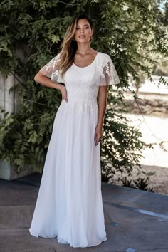 a woman wearing a white dress standing in front of some trees and bushes with her hands on her hips