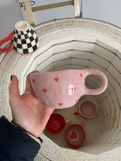 a hand is holding a pink cup with hearts painted on it and other items in the background