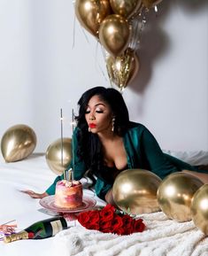 a woman sitting on a bed with a cake and candles in front of her, surrounded by balloons