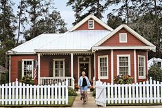 a woman walking down the sidewalk in front of a red house with white picket fence