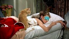 a woman laying in bed with two cats on top of her and one cat looking at her