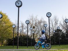 a young boy riding a bike in front of clocks on poles and trees with no leaves