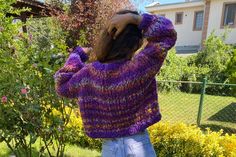 a woman wearing a purple sweater and jeans standing in front of some bushes with her hands on her head