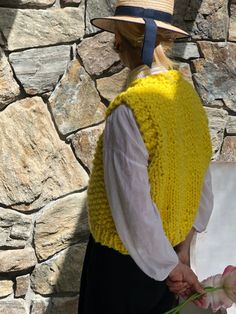 a woman wearing a yellow knitted vest and holding a flower in her hand while standing next to a stone wall