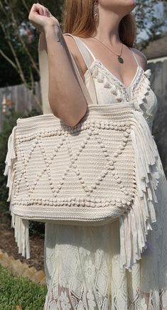 a woman in white dress carrying a crocheted bag with tassels on it