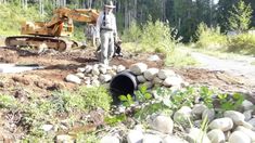 a man standing next to a pipe in the middle of a forest with a bulldozer behind him