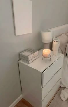 a night stand with two candles on it next to a white dresser and bed in a bedroom