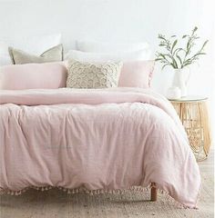 a bed with pink linens and pillows in a white room next to a wicker side table