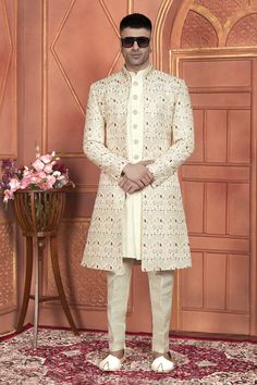 a man standing in front of a pink wall wearing a white sherwanit