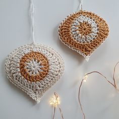 two crocheted ornaments hanging from strings on a white surface with lights around them