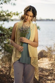 a woman is standing by the water with flowers in her hand and she is wearing a yellow knitted vest