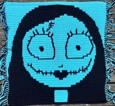 a crocheted square with a blue face and fringe around the edges on a wooden surface