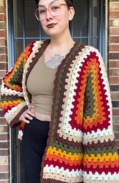 a woman standing in front of a brick building wearing a multicolored crocheted sweater