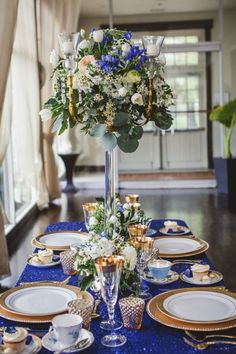 a table set with blue and gold plates, silverware and flowers in a tall vase