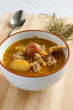 a bowl of soup with meat, potatoes and carrots next to a spoon on a cutting board