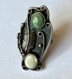 "an amazing and amazingly tall southwestern ring, artisan-made. top stone looks like a green turquoise, bottom one is a mother-of-pearl. beautiful feather and details, nice condition. marked with a stamping on the back. has a fair amount of patina but not much in the way of wear. mother-of-pearl shows some surface wear. size: 5.75 height of face: 1 and 15/16\" width of face: a bit over 13/16\" width of inner band: a bit over 1/16\"" Adjustable Green Opal Bohemian Ring, Green Adjustable Bohemian Opal Ring, Green Bohemian Opal Ring, Vintage Green Turquoise Ring Stamped 925, Vintage Adjustable Green Turquoise Ring, Bohemian Multi-stone Opal Ring, Bohemian Green Chrysoprase Ring, Green Bohemian Multi-stone Rings, Bohemian Green Multi-stone Rings