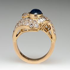 This elegant ring is centered with one (1), bezel set, oval cabochon cut natural sapphire. The sapphire is bordered with eight (8), channel set, tapered baguette cut diamonds and thirty-two (32), bead set, round brilliant cut diamonds. The ring measures 17.0mm at the top, rises 9.7mm above the finger, tapering to 3.8mm wide and 2.0mm thick at the base of the shank. This ring is currently a size 6. Elegant Gold Sapphire Ring With Oval Cabochon, Elegant Sapphire Diamond Ring Channel Set, Formal Domed Cabochon Sapphire Ring, Luxury Yellow Gold Sapphire Ring Channel Set, Elegant Domed Sapphire Ring In Yellow Gold, Elegant Domed Yellow Gold Sapphire Ring, Luxury Oval Sapphire Ring With Polished Finish, Formal Baguette Cut Sapphire Ring Channel Set, Formal Domed Blue Sapphire Ring