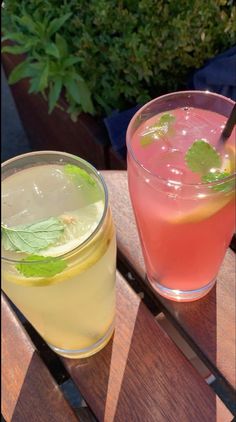 two drinks sitting next to each other on top of a wooden table with plants in the background