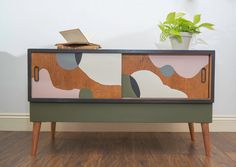 a wooden cabinet with an abstract design painted on the front and sides, sitting in a room next to a potted plant