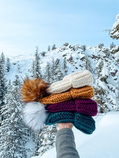 a person's hand holding several knitted scarves in front of snow - covered trees