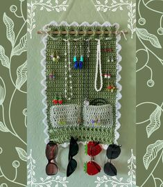 a crocheted wall hanging with sunglasses and other items on it, in front of a green floral background