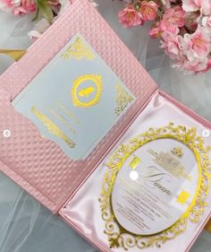 an open pink and gold wedding card in a box next to flowers on a table