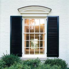 an open window with black shutters on a white brick wall and shrubbery in the foreground