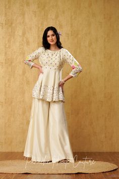 "Introducing our stunning Ivory Peplum Top with Sharara Pant, adorned with mesmerizing mirror and resham embroidery, crafted from luxurious Chanderi silk fabric." The Peplum Top is exquisitely designed to flatter the silhouette, with a chic and contemporary look. The intricate mirror and resham embroidery on the top adds a touch of elegance, creating a stunning visual effect. The top features a round neckline, complemented with a sleeveless design, which makes it perfect for any special occasion Peplum With Skirt Indian, Peplum Top Outfits Indian With Sharara, Peplone Design, Chanderi Palazzo Set With Gota Work For Reception, Bollywood Style Chanderi Palazzo Set In Off White, Off White Palazzo Set With Zari Work For Festivals, Off White Sharara For Festivals With Straight Kurta, Bollywood Style Off White Chanderi Palazzo Set, Off White Sharara With Straight Kurta For Festivals