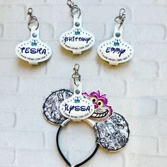 three mickey mouse ears with tags attached to them on a brick wall background, one has a name tag and the other has an image of a cartoon character's head