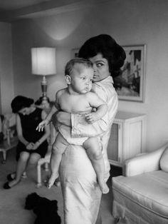 a woman holding a baby while standing in a living room with other people behind her