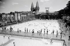 many people are standing around in the water at an outdoor swimming pool with large buildings in the background