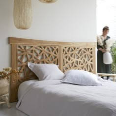 a bed with white sheets and pillows next to a woman standing in the window behind it