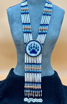 Bone hairpipes and crystal beads are often seen as part of traditional Native American dancers.  This modified breastplate  has been created to proudly proclaim that this is a good day to be Indigenous . It is designed to be worn not only in the dance circle, but in mainstream life, showing your Native pride and fashion sense.   The necklace is 10 vertical strands of bone hairpipes, nickel beads and 8mm sparking, faceted, fire-polished glass crystal beads in the colors of navy blue, turquoise blue and white frost The centerpiece is a beaded rosette of a Bear paw in the same blue colors on a white background. At the bottom of breastplate, pewter and rhinestone pendants  add their music to your movements. The breastplate comes with matching earrings on hypoallergenic lever back wires featuri Native American Medallions, Powwow Regalia Jingle Dress, Native Medallion, Indigenous Jewelry, Native American Dance, American Dance, Powwow Regalia, Creative Fabric, Jingle Dress