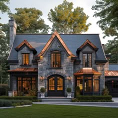 a large stone house with lots of windows and lights on the front door, surrounded by greenery